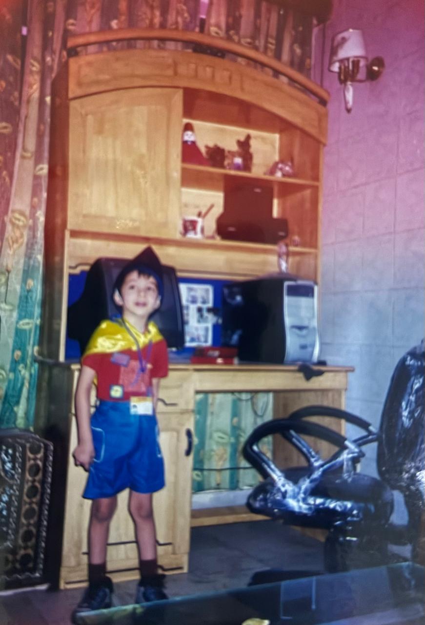 A young Naman standing in front of a CRT computer at home, the cabinet behind him and the curtains drawn